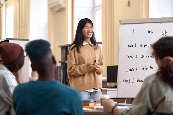 A smiling Asian woman with shoulder-length black hair stands before three peers with a black marker in hand. She wears a white shirt beneath a tan sweater with blue jeans. To her right is a whiteboard with syllables of a language written out and English words – I want, I need, I can, I think – written next to them. Her peers, blurred but visible, are facing her and away from the camera. They are in a brightly lit building space.